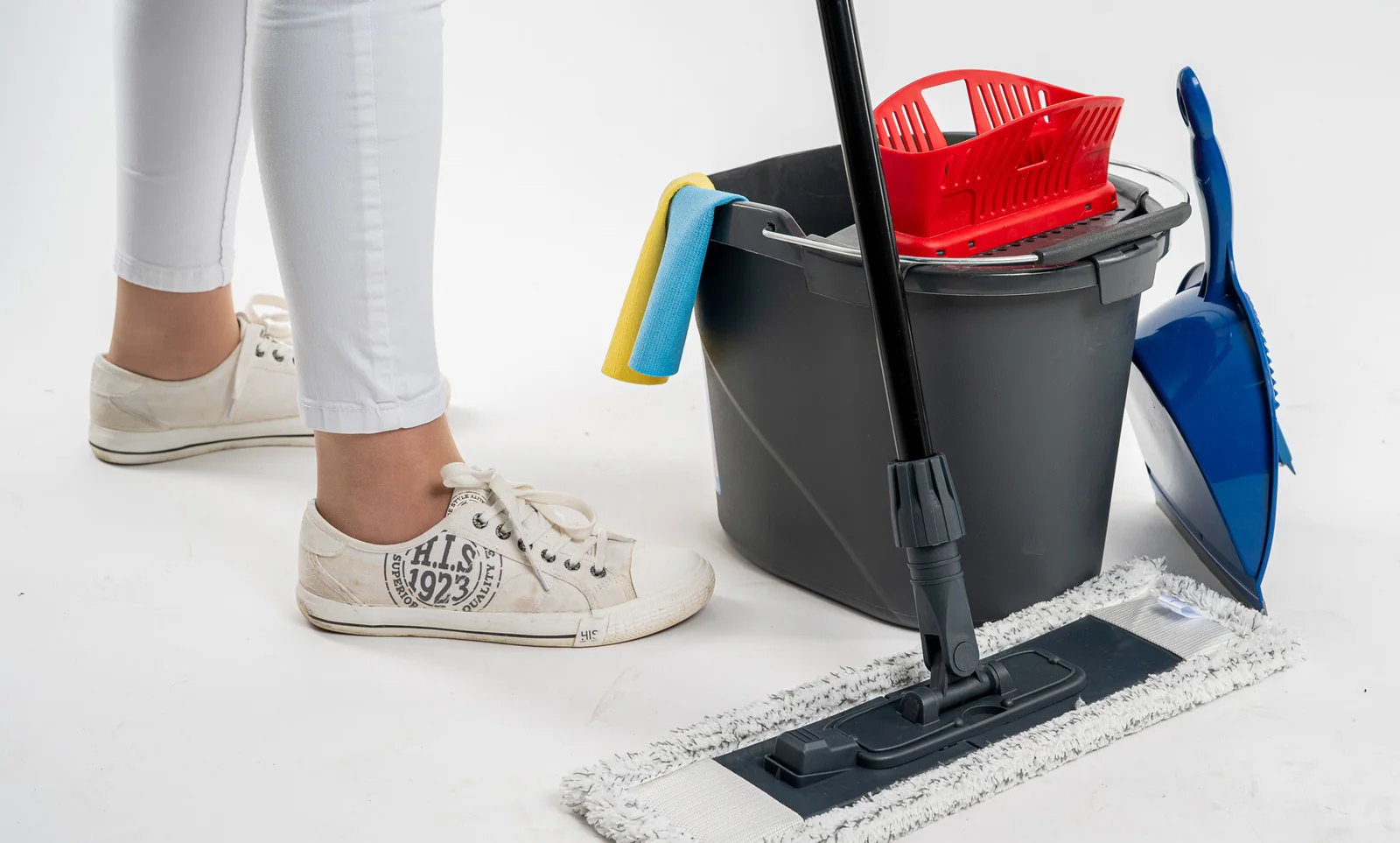 Eine Hauswirtschaftskraft in weißer Hose steht neben Wischeimer, Bodenwischer und Kehrschaufel